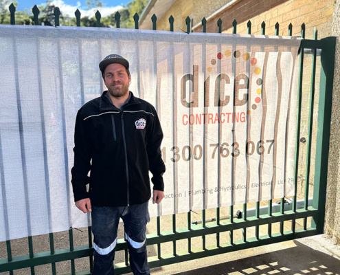 Dice Contracting worker standing in front of a fence with company banner displaying contact details.